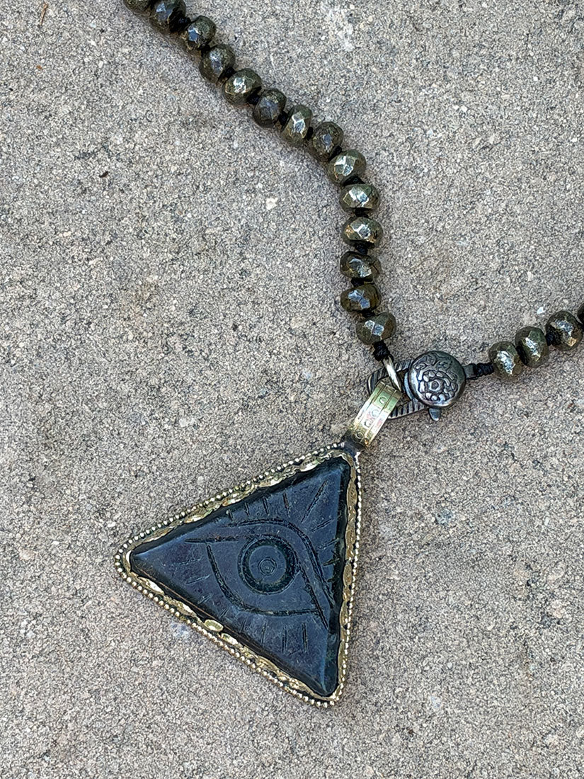 A close-up of a beaded necklace with faceted pyrite beads and a triangular pendant featuring an eye motif in dark blue-gray stone with a silver-toned border, laid on a textured concrete surface.
