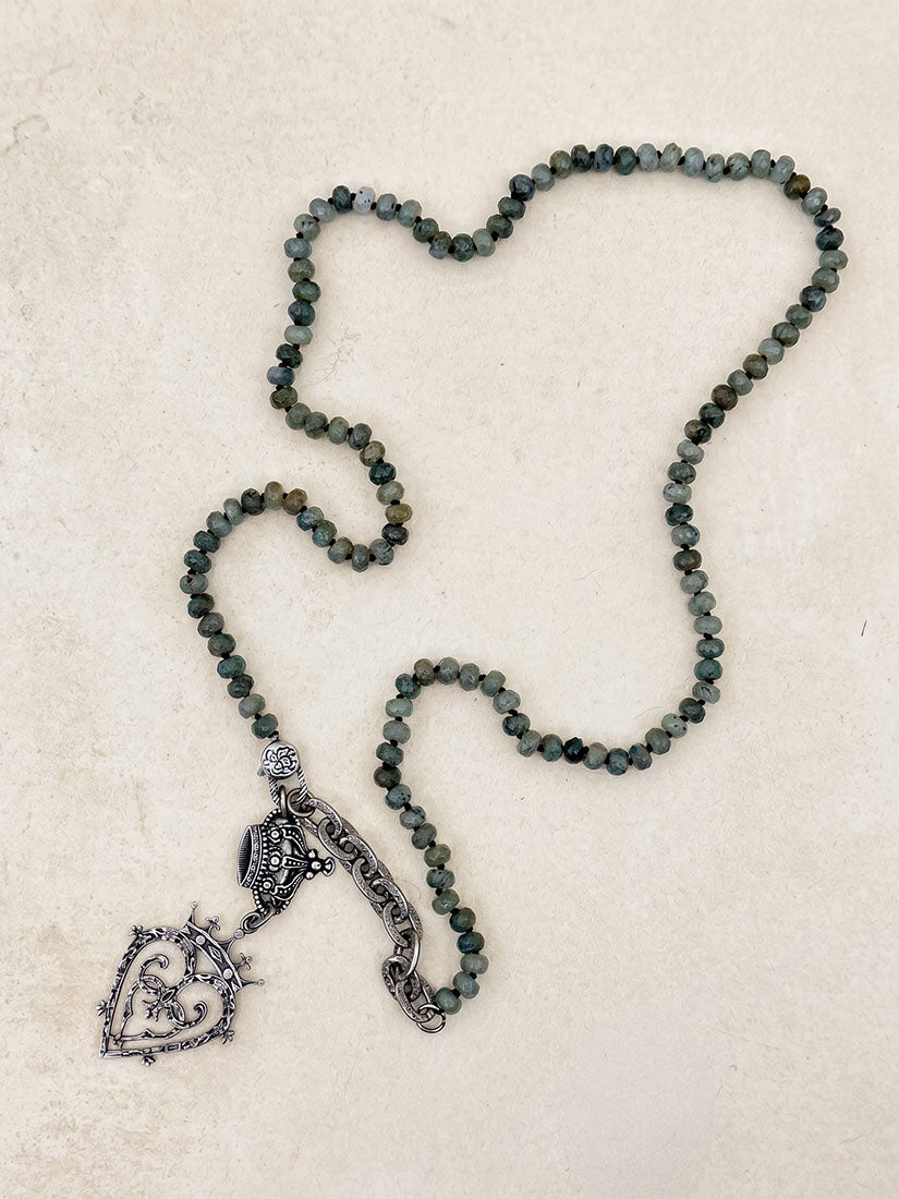 A beaded necklace with dark gray and green labradorite faceted beads, featuring a silver-toned filigree crown pendant and a heart-shaped charm, displayed on a textured beige surface.