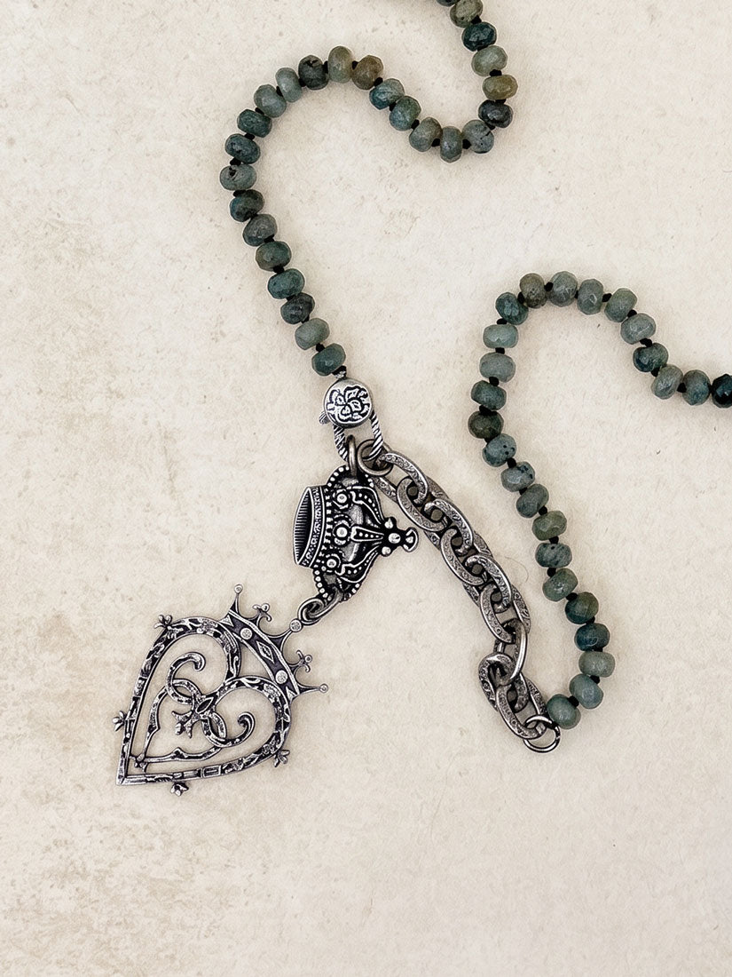 A beaded necklace with dark gray-green round beads and an ornate silver-tone pendant featuring a heart-shaped filigree design, laid flat on a light beige textured surface.