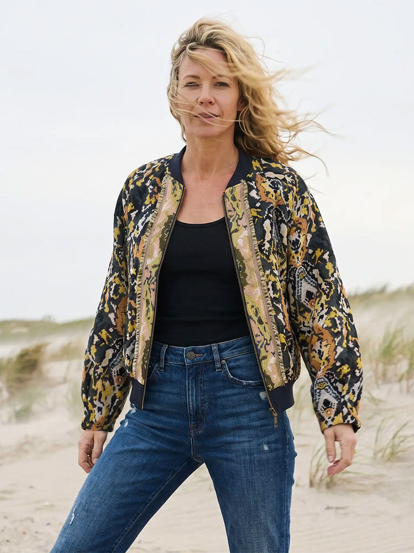 A woman standing on a sandy beach wearing a black bomber jacket with a gold, beige, and yellow abstract pattern, a black tank top, and blue jeans.