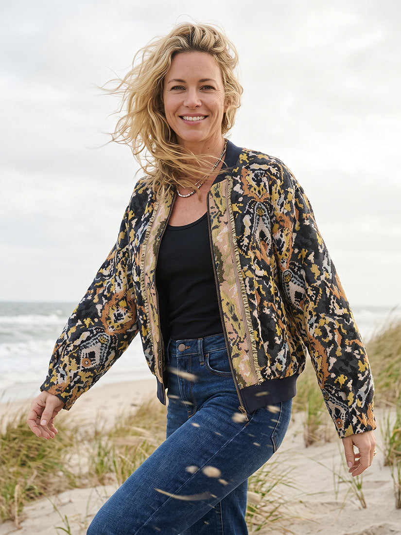 A woman stands on a sandy beach wearing a black bomber jacket with a detailed gold, beige, and brown tribal-style print, a black tank top, and blue jeans.