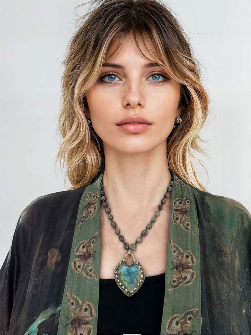A woman with blonde-highlighted hair wearing a green butterfly-patterned cardigan over a black top, featuring a bronze-toned beaded necklace with a large blue-green heart-shaped pendant.