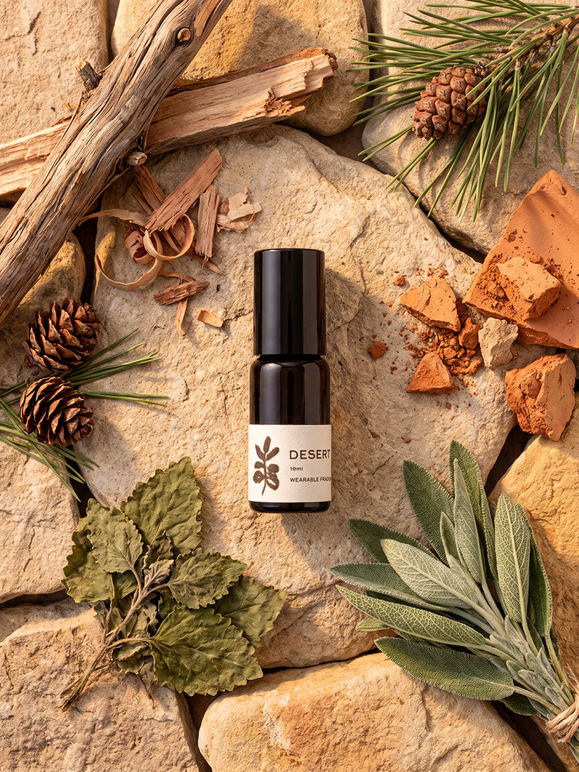 A small dark amber bottle with a black cap and a cream label reading 'DESERT' and 'MEXICO' is centered on a flat lay of natural stones, pine needles, pinecones, dried leaves, bark, and earthy props.