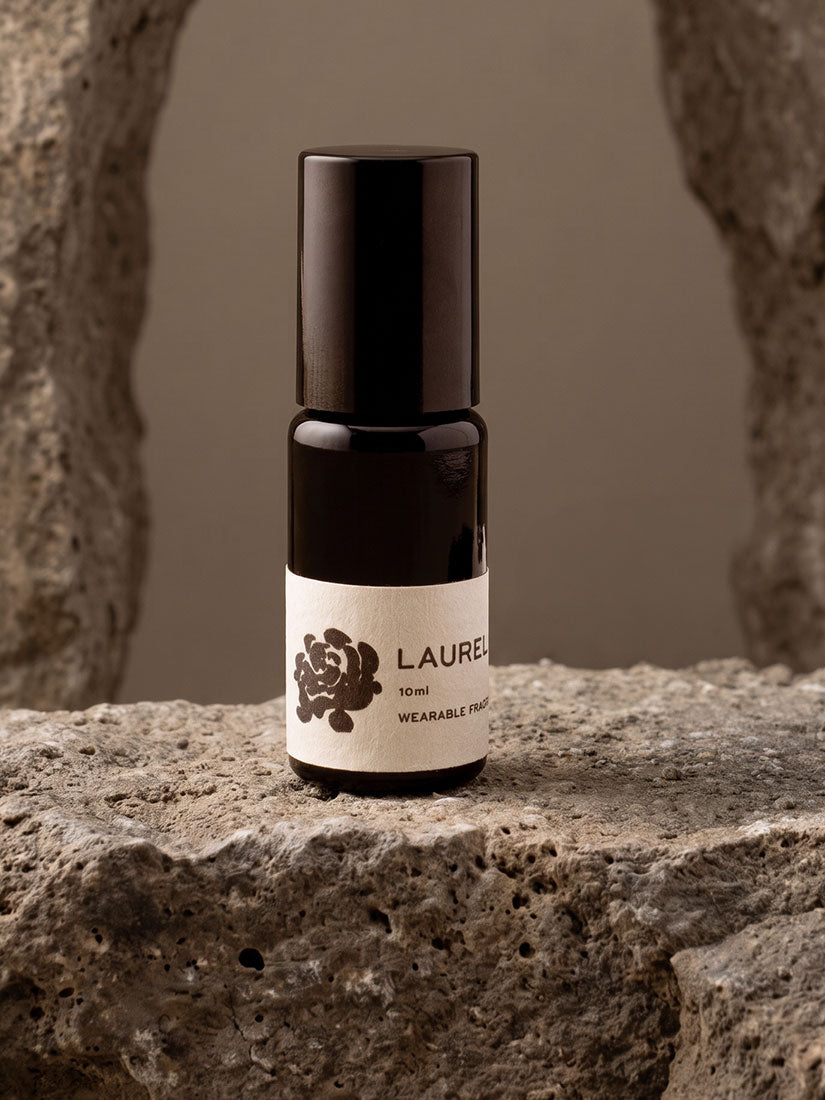 A 10 ml bottle of Laurence Wearable Scents wearable perfume, featuring a black cap and a cream-colored label with a dark botanical illustration, set on a textured stone surface.