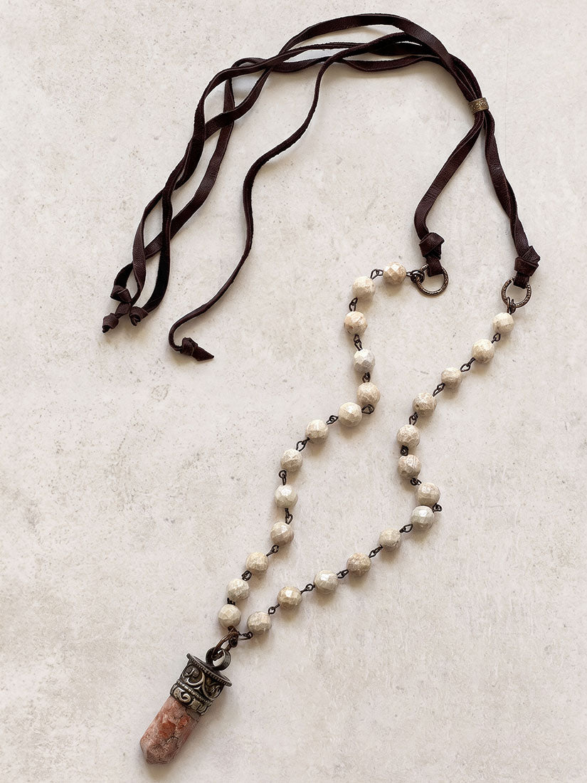 A bohemian-style beaded necklace featuring cream-colored seed beads, dark brown leather ties, and a carved orange stone pendant in a silver-toned metal cap, displayed on a textured light beige background.