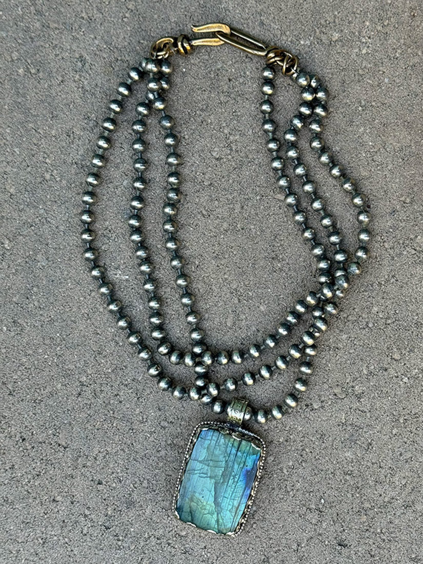 A multi-strand necklace featuring dark gray metallic beads and a rectangular labradorite pendant with blue and green iridescence, set in silver-toned metal on a textured gray background.