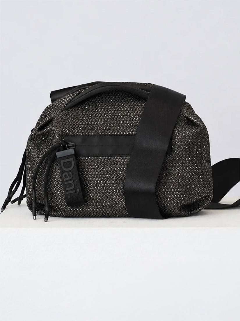 A black woven crossbody bag with a wide black strap, a black hanging tag reading 'Dant', and black fringe detail, photographed against a plain light gray studio background.
