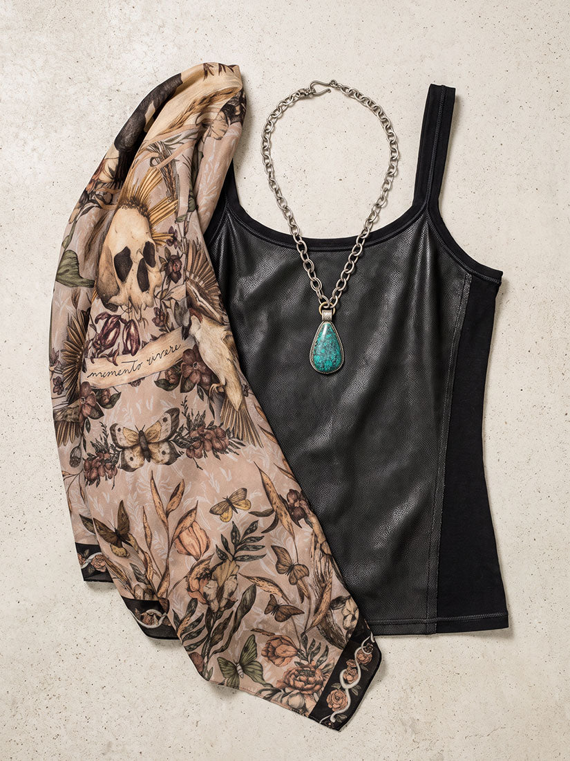 A styled flat-lay featuring a black faux-leather tank top, a silver chain necklace with a turquoise pendant, and a beige patterned scarf with a skull and floral print, arranged on a textured light beige background.