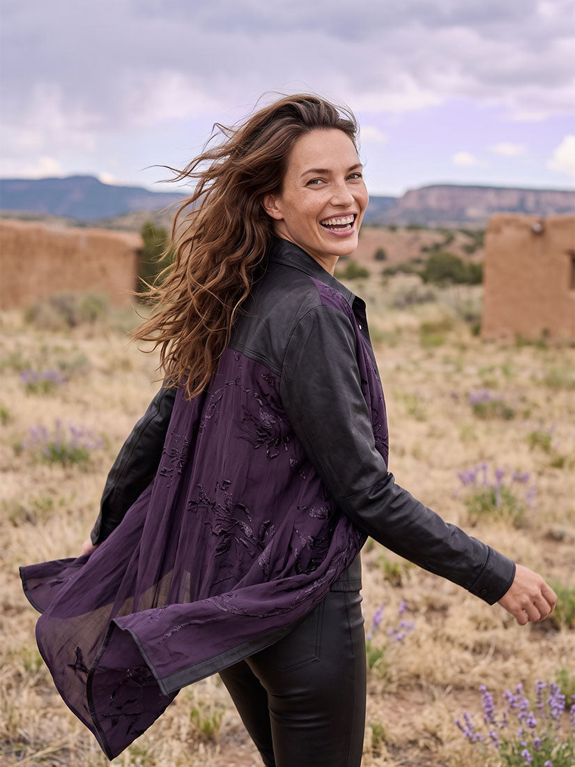 A woman outdoors in a desert landscape wearing a dark purple embroidered tunic-style jacket over a black leather jacket and black pants, smiling while looking back over her shoulder.
