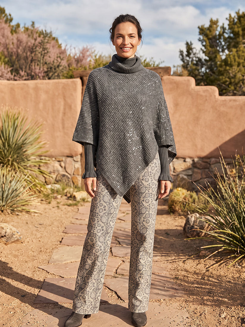 A woman stands outdoors in a desert courtyard wearing a dark gray textured turtleneck poncho and light gray patterned wide-leg pants.