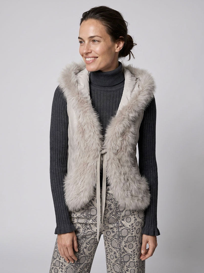 A woman wearing a light gray faux fur vest over a dark gray ribbed turtleneck sweater and gray patterned pants, photographed against a neutral light gray studio background.