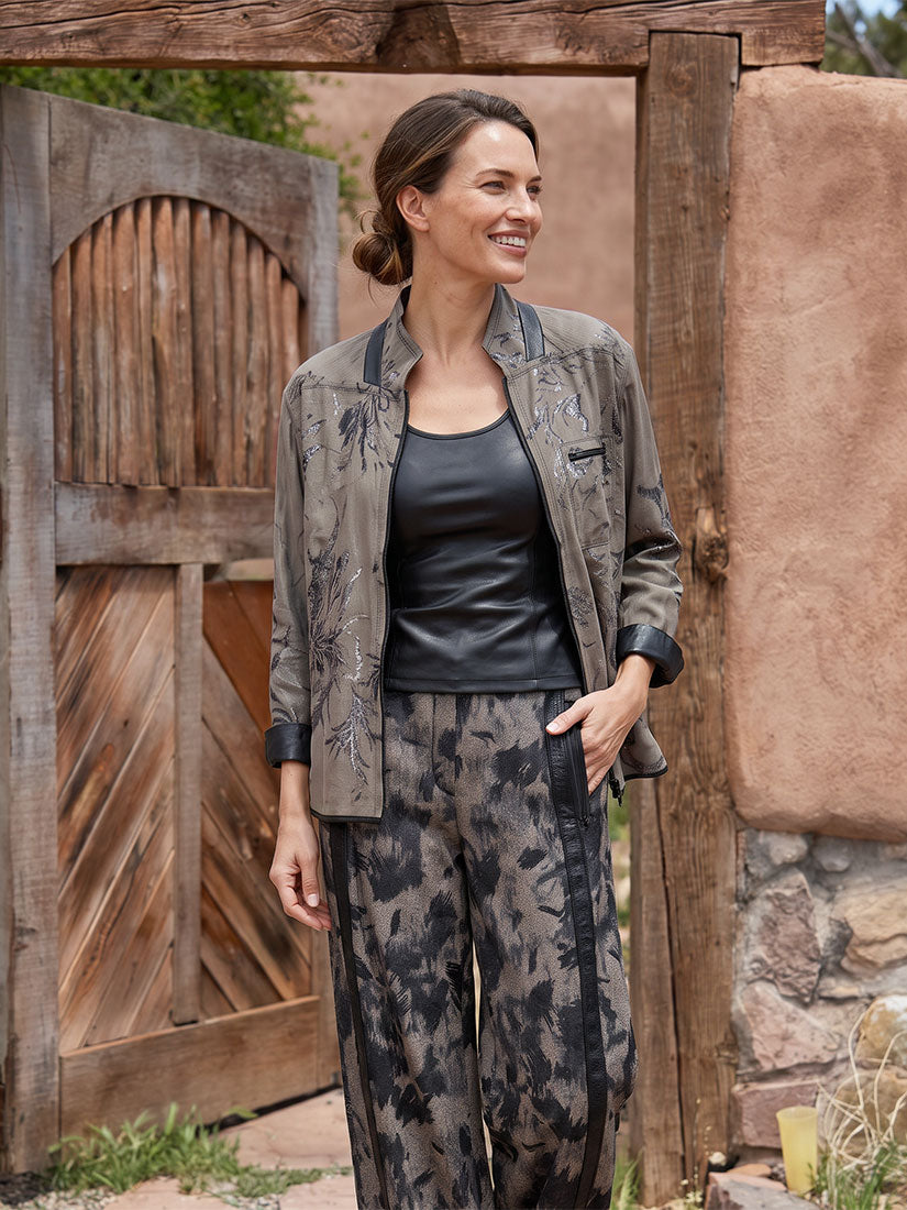 A woman stands outdoors in front of a rustic wooden gate and adobe wall, wearing a matching two-piece outfit with a taupe and black botanical print jacket and wide-leg pants over a black top.