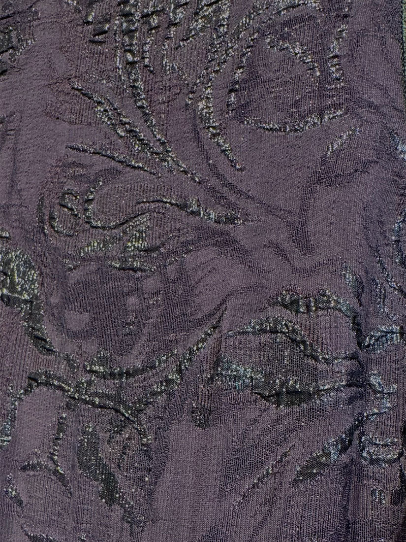 A close-up of a textured fabric with a floral damask pattern in a muted plum purple with metallic silver highlights.