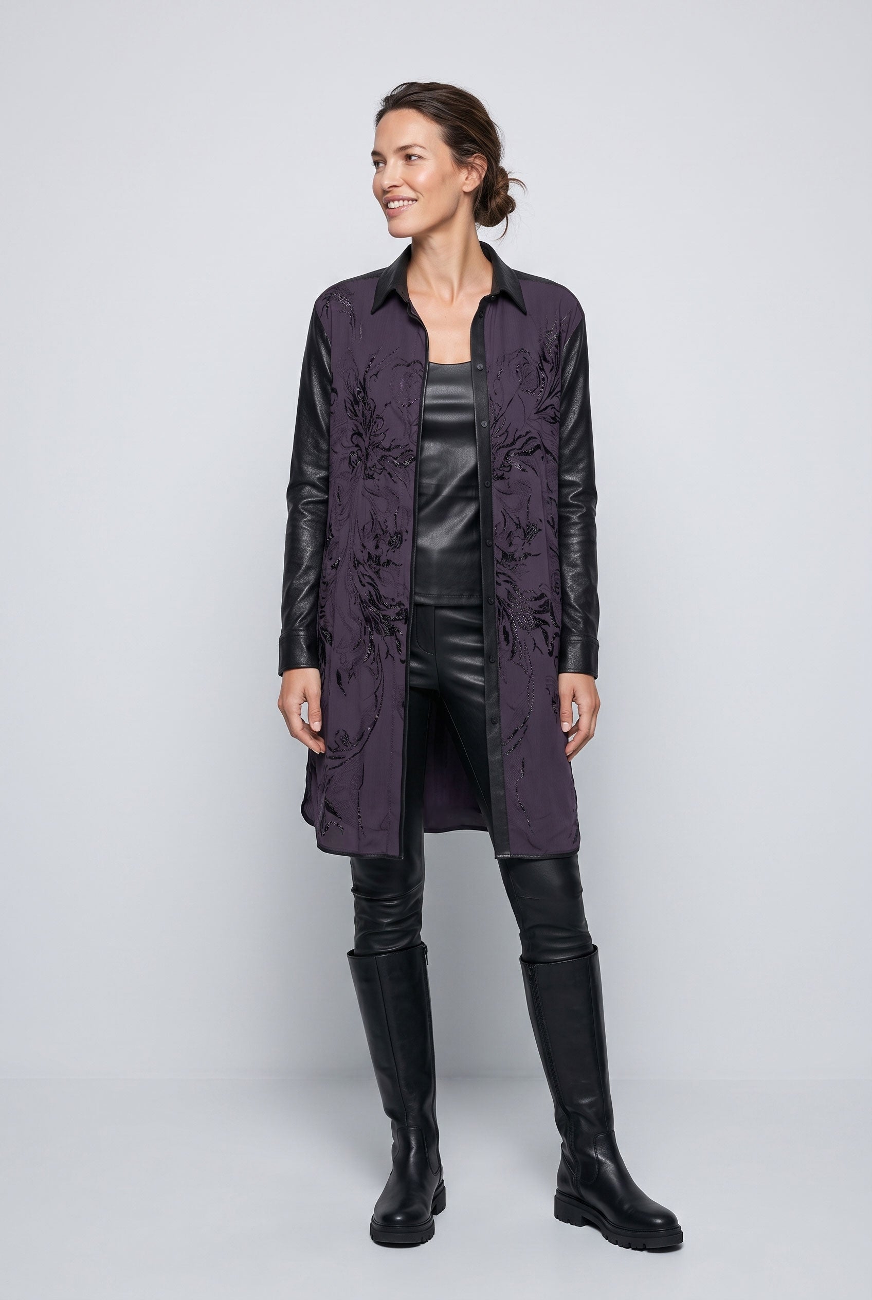 A woman models a longline open-front coat with dark purple floral jacquard fabric and black faux-leather sleeves, styled with a black leather top, black leather pants, and black knee-high boots against a light gray studio background.