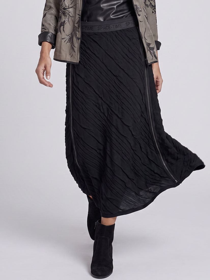 A model wearing a black pleated midi skirt with side zippers, styled with a black leather top, a brown patterned jacket, and black suede ankle boots against a light gray studio background.