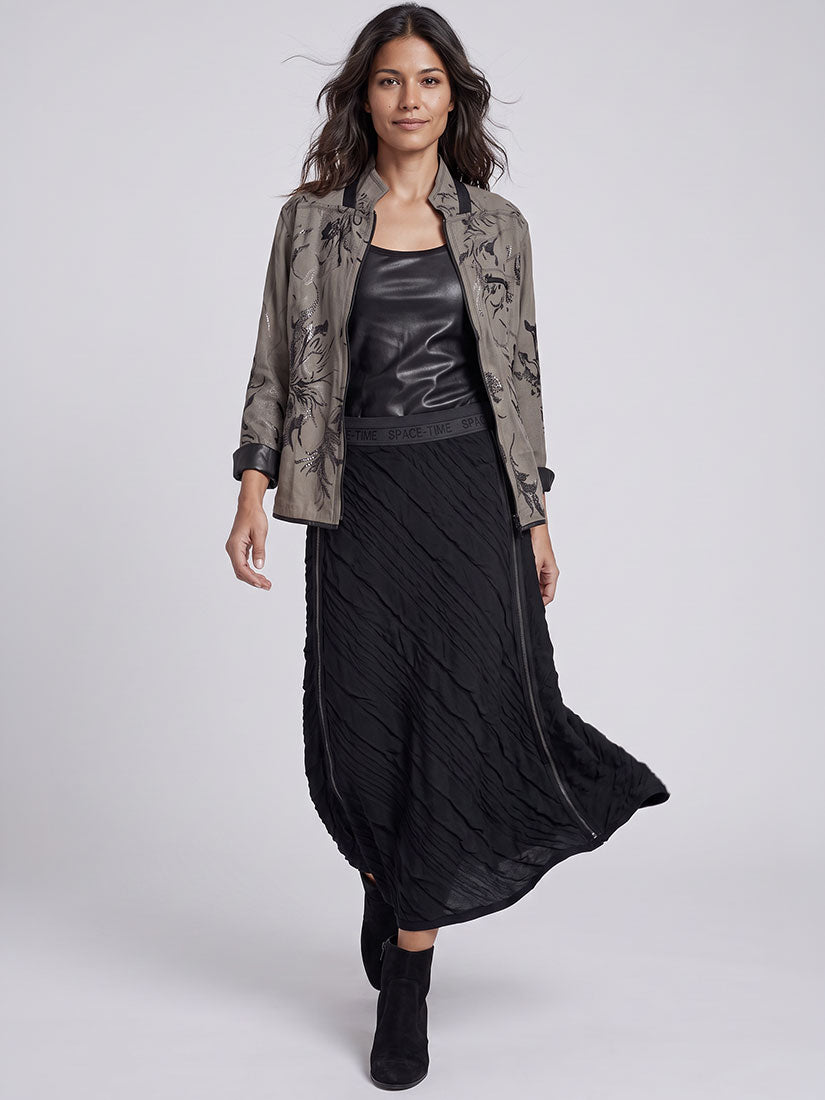 A model walks against a light gray studio background wearing a black pleated midi skirt, a black faux-leather top, and a textured taupe-brown jacket with black floral embroidery.