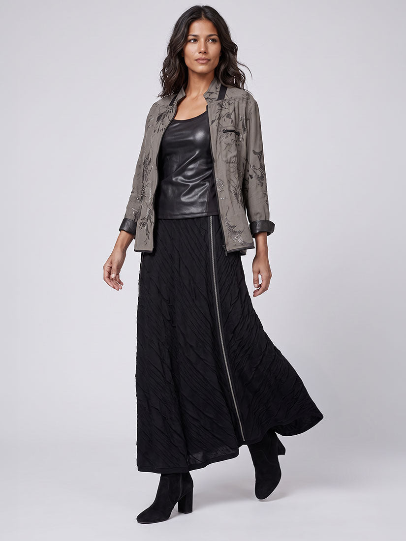 A model is shown wearing a styled outfit consisting of a textured black leather top, a long black quilted skirt with a front zipper, a distressed gray-brown jacket, and black ankle boots, set against a plain light gray studio background.