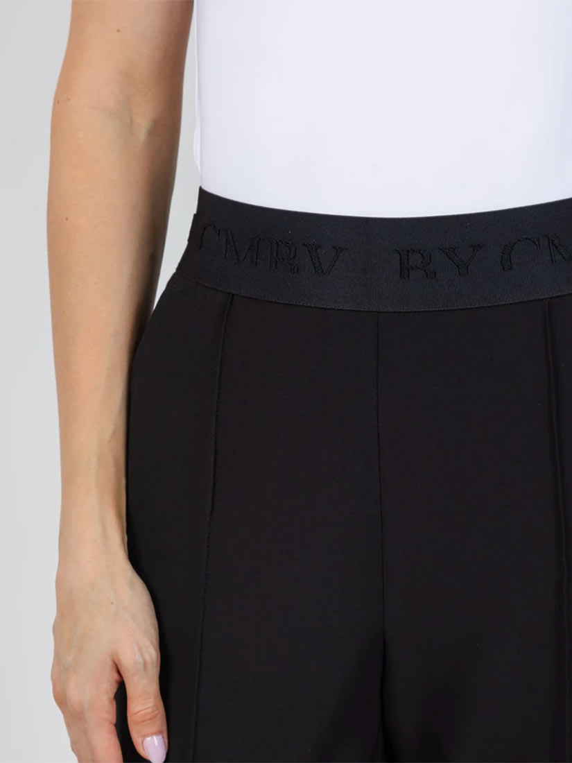 Close-up of a model wearing black trousers with a wide black elastic waistband featuring embossed brand lettering, paired with a white top against a light gray background.
