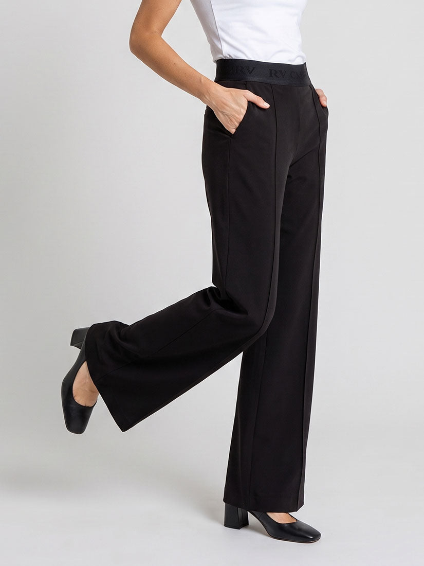 A model wearing black wide-leg trousers with a satin waistband, styled with a white top and black heels, shown from the waist down against a light gray studio background.