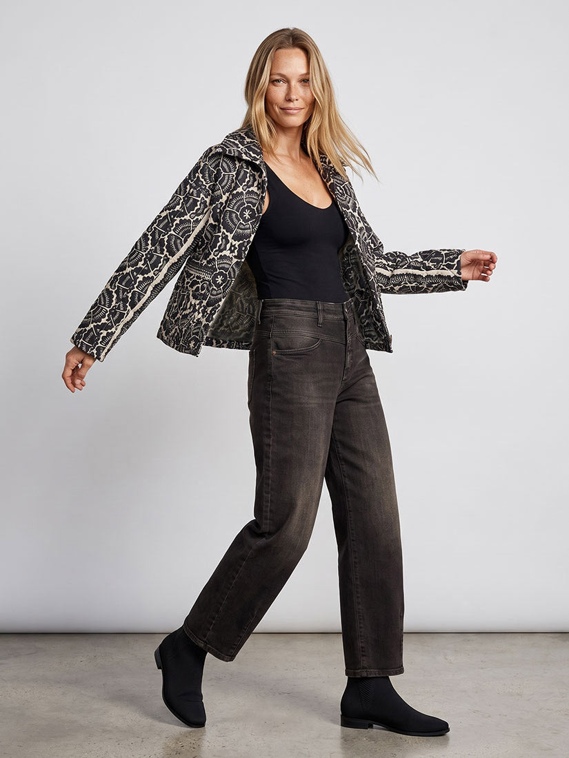 A woman modeling a black and beige floral patterned jacket over a black tank top and dark gray wide-leg jeans, standing against a light gray studio background.