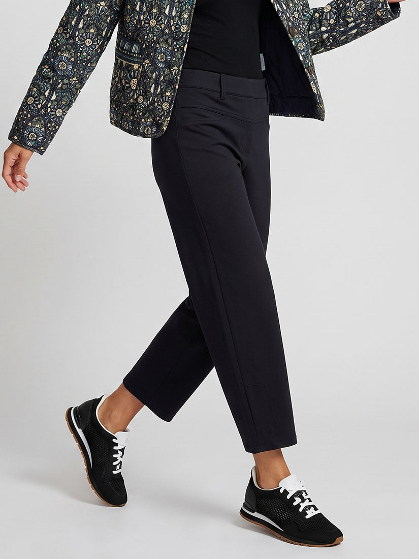 A person wearing slim black cropped trousers, a black top, a patterned blazer, and black sneakers with white laces, photographed against a light gray studio background.