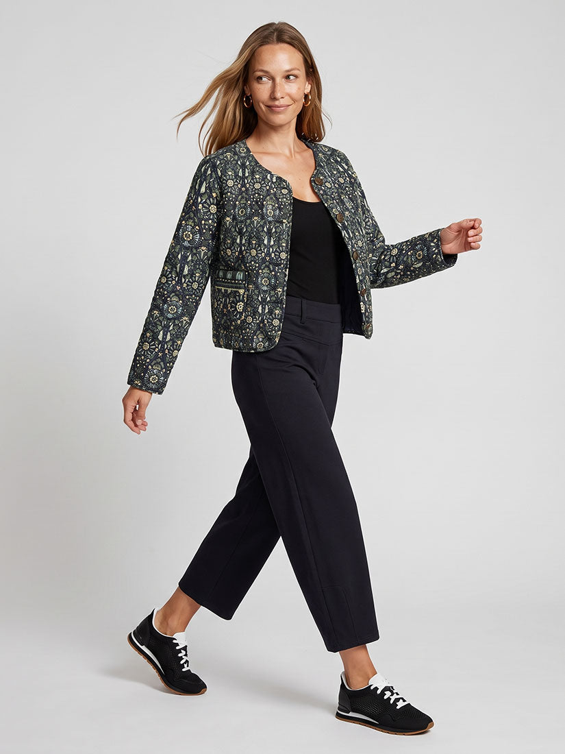 A woman modeling a cropped, long-sleeve patterned jacket with a black top, black cropped trousers, and black sneakers against a light gray studio background.