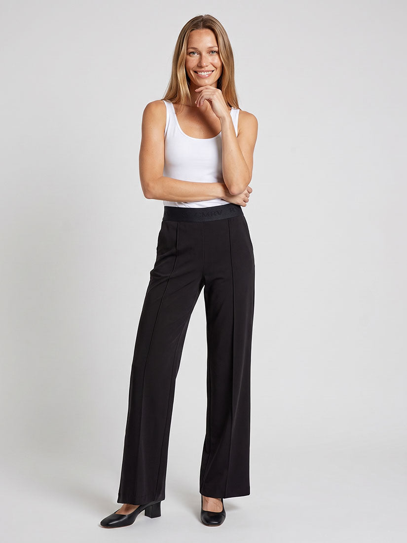 A woman modeling black wide-leg trousers with a contrasting black waistband, paired with a white ribbed tank top and black heels, against a light gray studio background.