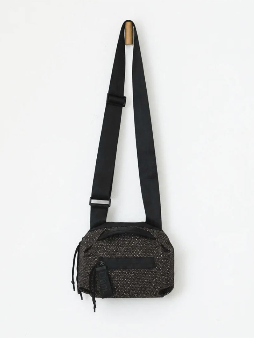 A black textured shoulder bag with a wide adjustable black webbing strap, hanging from a wooden peg against a plain white wall.