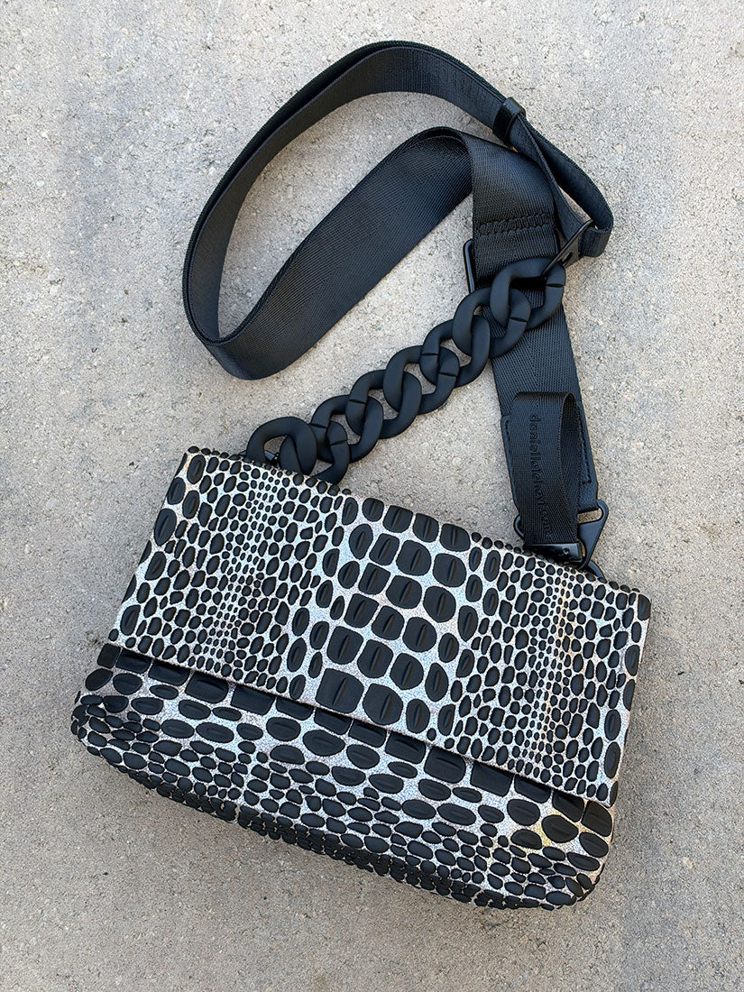A black and silver tokyo snakeskin-patterned shoulder bag with a wide black strap from daniella lehavi, laid flat on a light gray concrete surface.