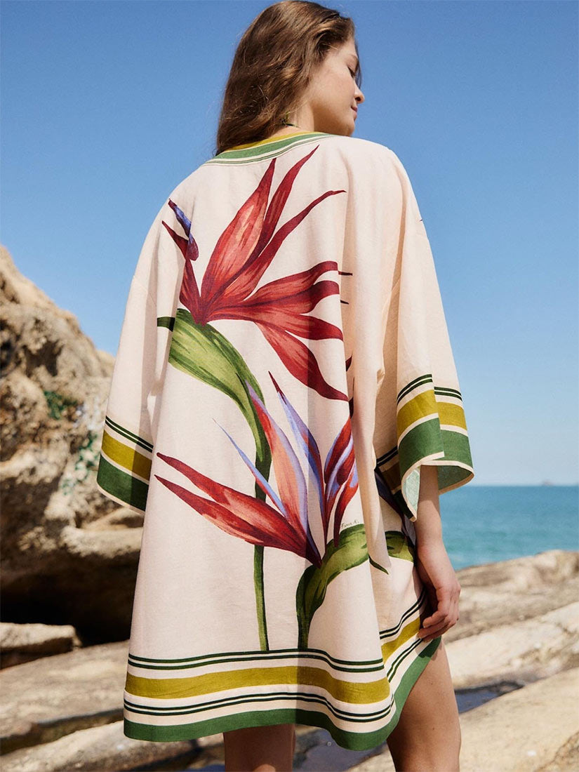 A woman stands on a rocky beach wearing a cream-colored kaftan cover-up with a large red and green tropical bird of paradise floral print and striped trim.