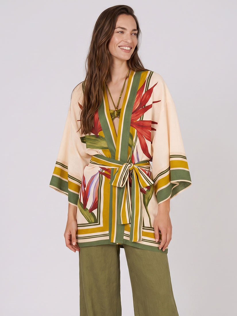 A woman modeling a cream-colored wrap kimono with a green, yellow, and red tropical floral print, paired with olive green wide-leg pants, against a light gray studio background.