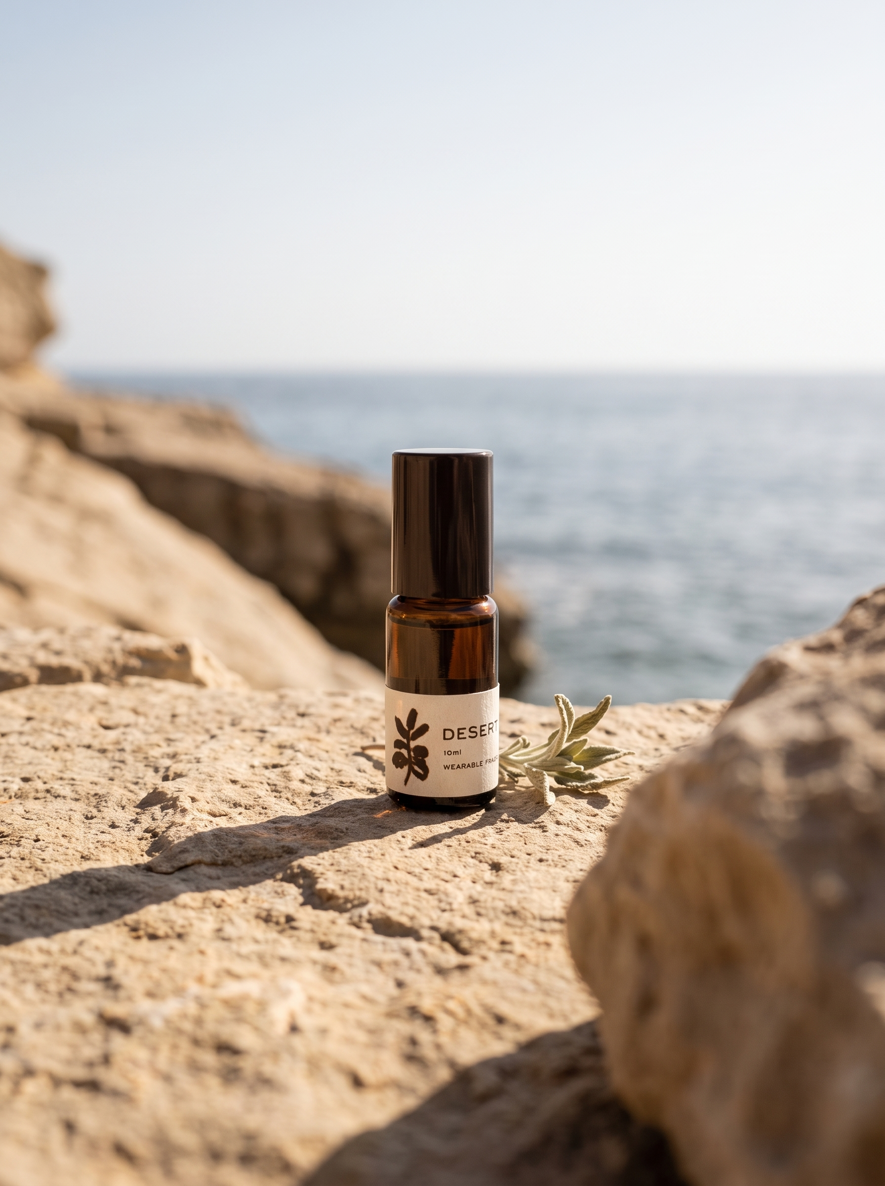 A small amber glass bottle with a black cap and a cream label reading DESIGNS by JENNIE stands on a sunlit rocky beach with the sea blurred in the background.