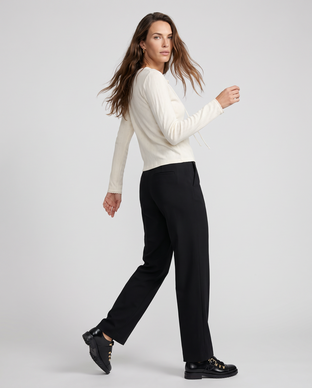A woman modeling a cream ribbed long-sleeve top and black wide-leg trousers, paired with black leather shoes, against a light gray studio background.