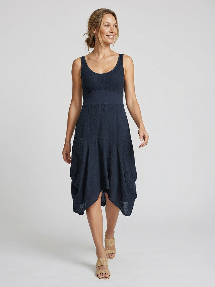A woman modeling a sleeveless navy blue midi dress with a scoop neckline, textured fabric, and an asymmetrical draped hem, paired with beige strappy sandals against a light gray studio background.