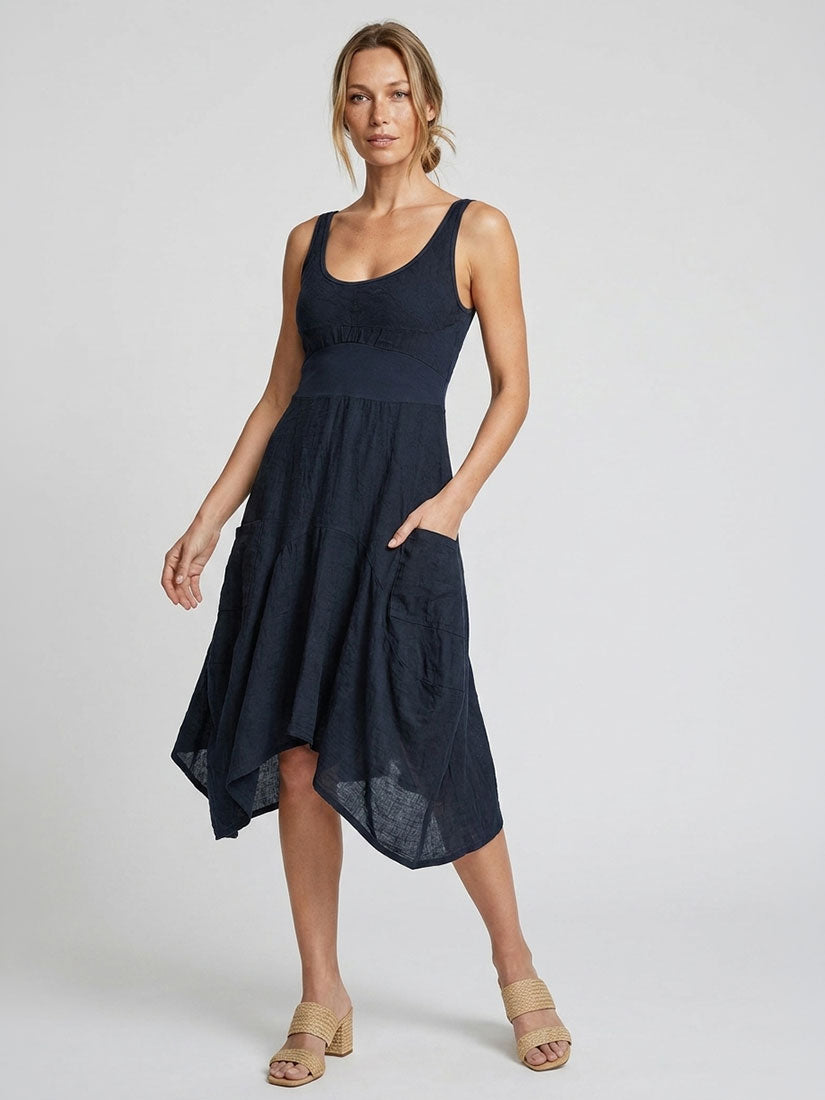 A woman modeling a sleeveless navy blue midi dress with a smocked bodice, tiered skirt, and side pockets, paired with beige heeled sandals against a light gray studio background.