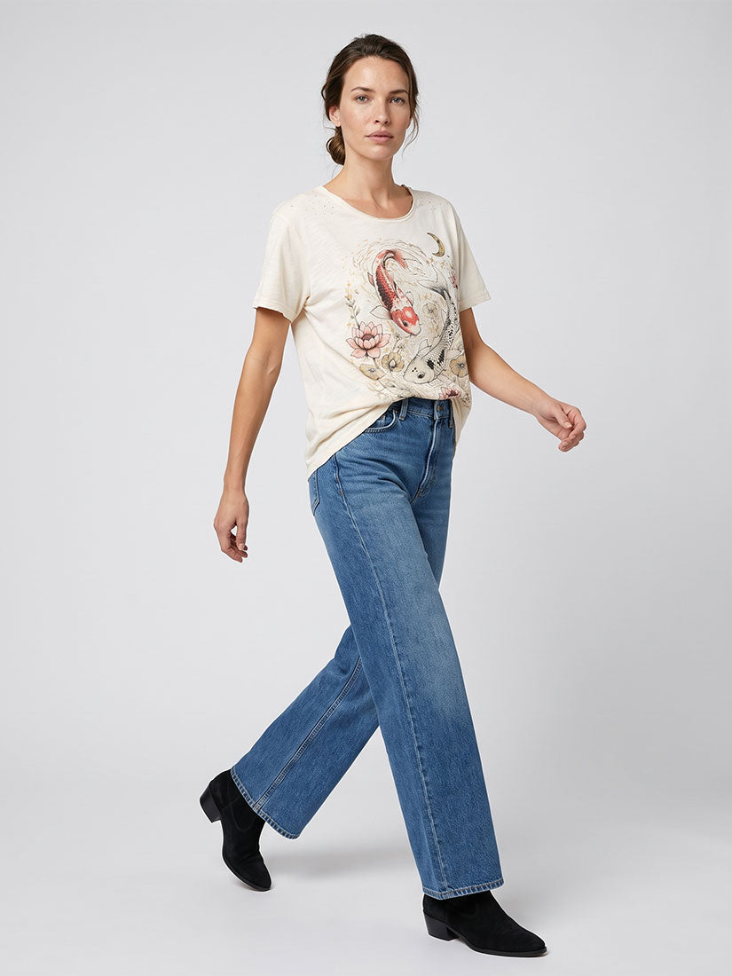 A woman modeling a cream graphic t-shirt with a koi fish print, paired with blue jeans and black ankle boots, walking against a light gray studio background.