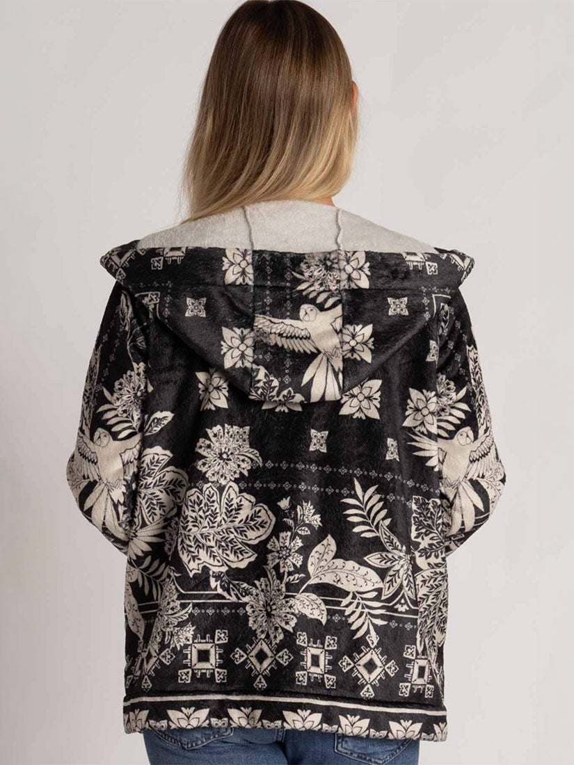 A woman viewed from behind wearing a black hooded jacket with a detailed white and gray bird and floral pattern, paired with blue jeans against a plain light gray background.