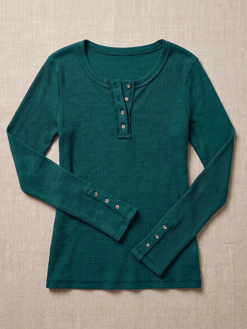 A dark teal green ribbed long-sleeve henley shirt with button details on the collar and cuffs, laid flat on a textured beige fabric background.