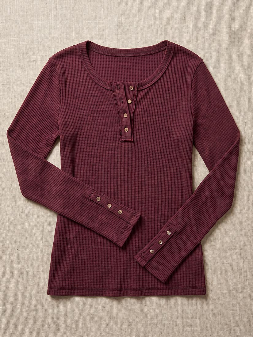 A long-sleeve waffle-knit henley shirt in deep burgundy, laid flat on a textured beige fabric background. The shirt features a three-button placket on the chest and three-button cuffs.