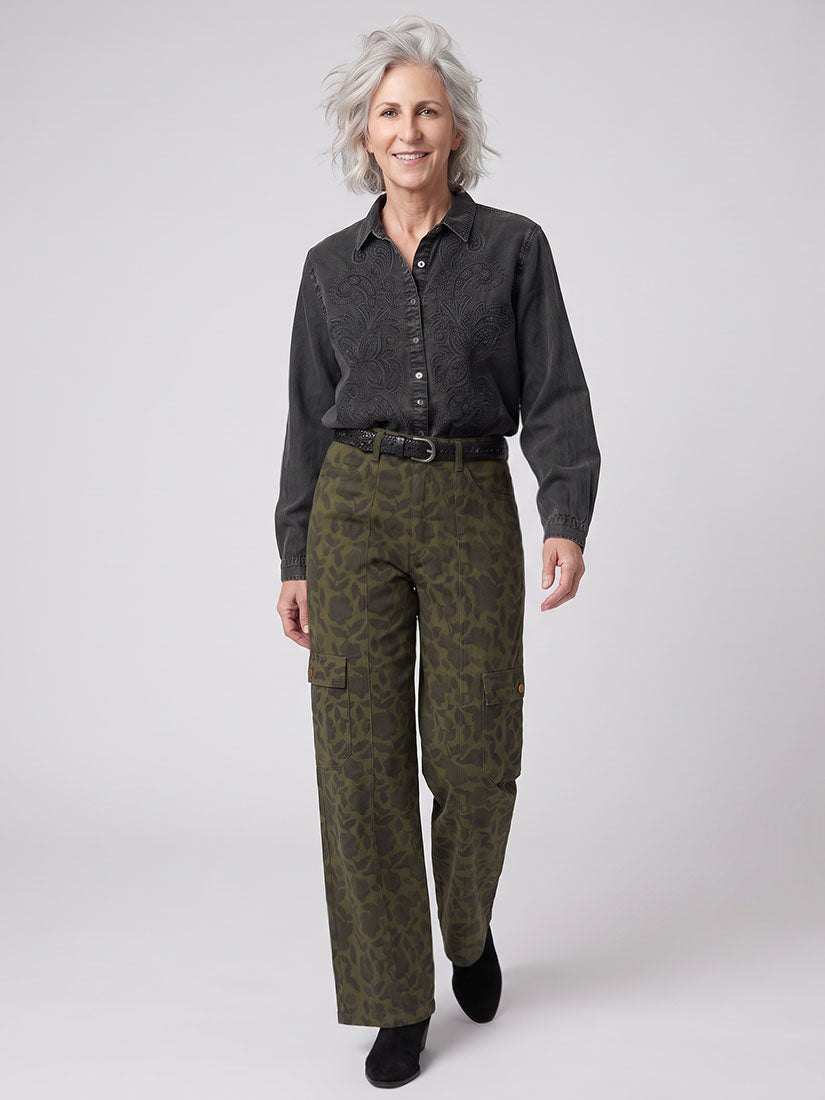 An older woman modeling a dark charcoal gray long-sleeve button-up shirt and olive green floral patterned cargo pants against a light gray studio background.