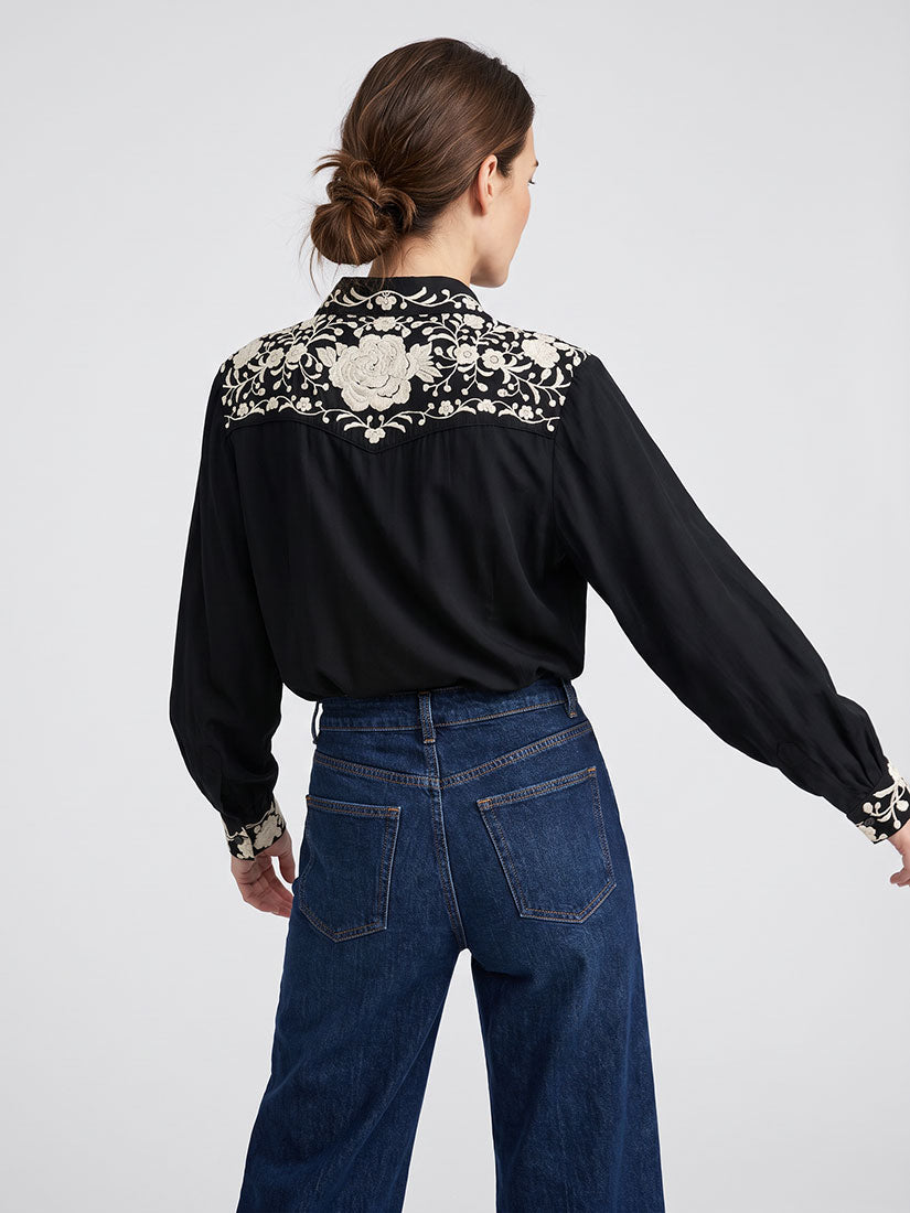 A woman shown from the back wearing a black long-sleeve blouse with a large beige floral embroidery panel across the shoulders and dark blue wide-leg jeans, against a light gray studio background.