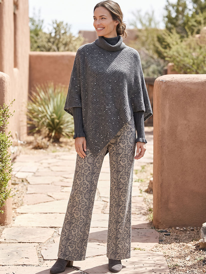 A woman stands outdoors in a desert-style setting wearing a dark gray turtleneck poncho with subtle sequin details and matching gray patterned wide-leg pants.