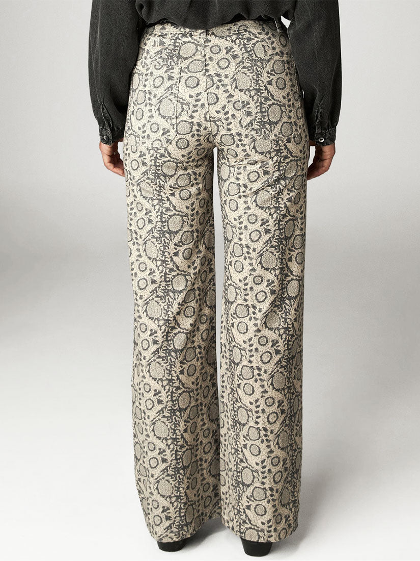 A person shown from the back wearing high-waisted, wide-leg trousers with an allover abstract circular pattern in beige and gray, paired with a dark long-sleeve top.