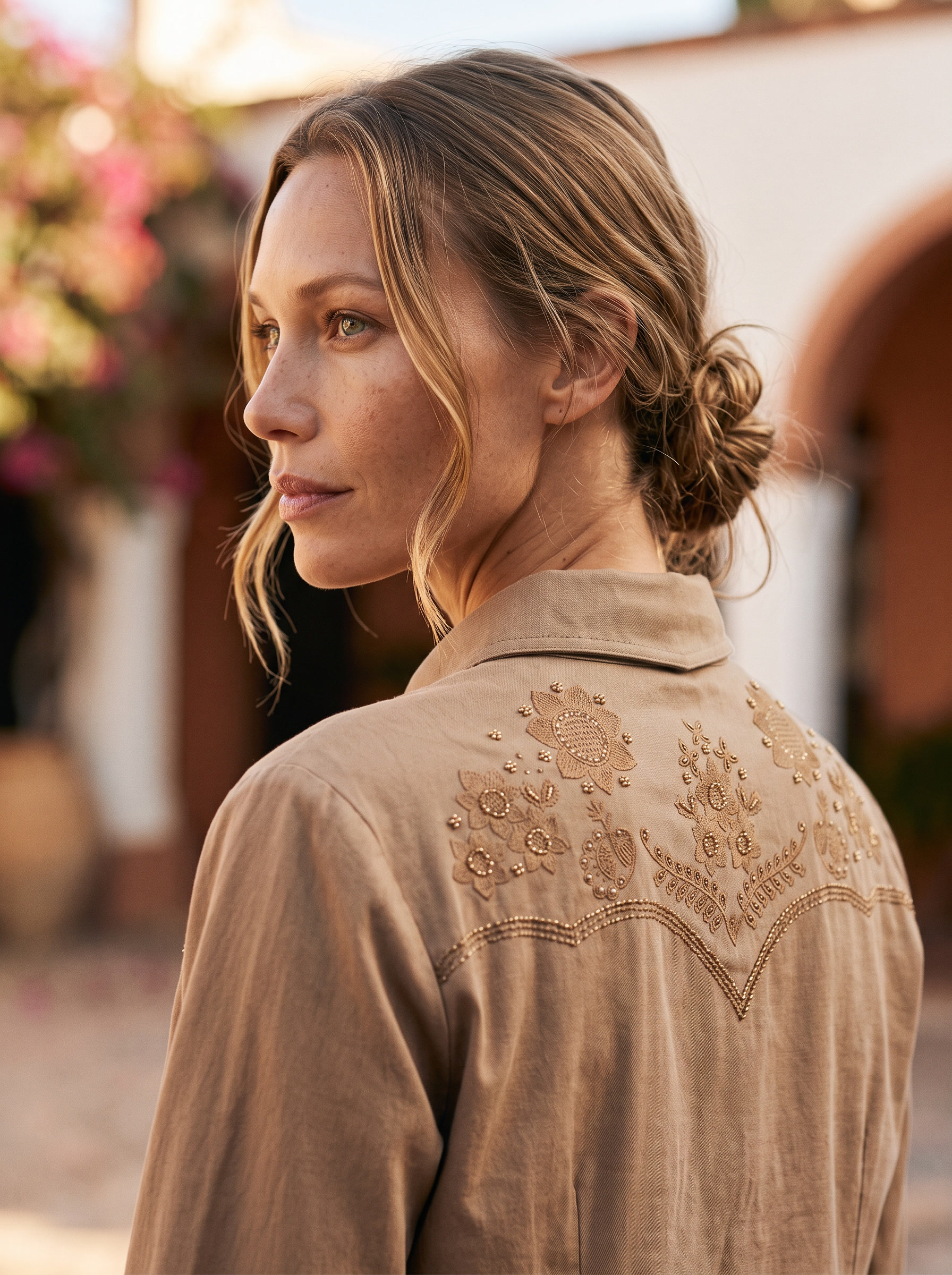 A woman shown from the back wearing a tan embroidered long-sleeve shirt in a warm, sunlit outdoor setting.