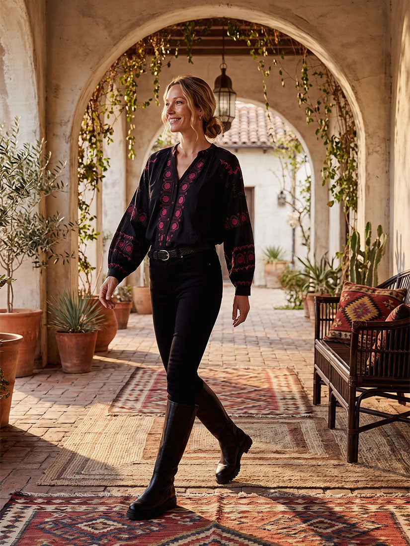 A woman walks through a sunlit courtyard with arches, plants, and patterned rugs, wearing a dark long-sleeve blouse with red floral embroidery, black pants, a black belt, and black knee-high boots.