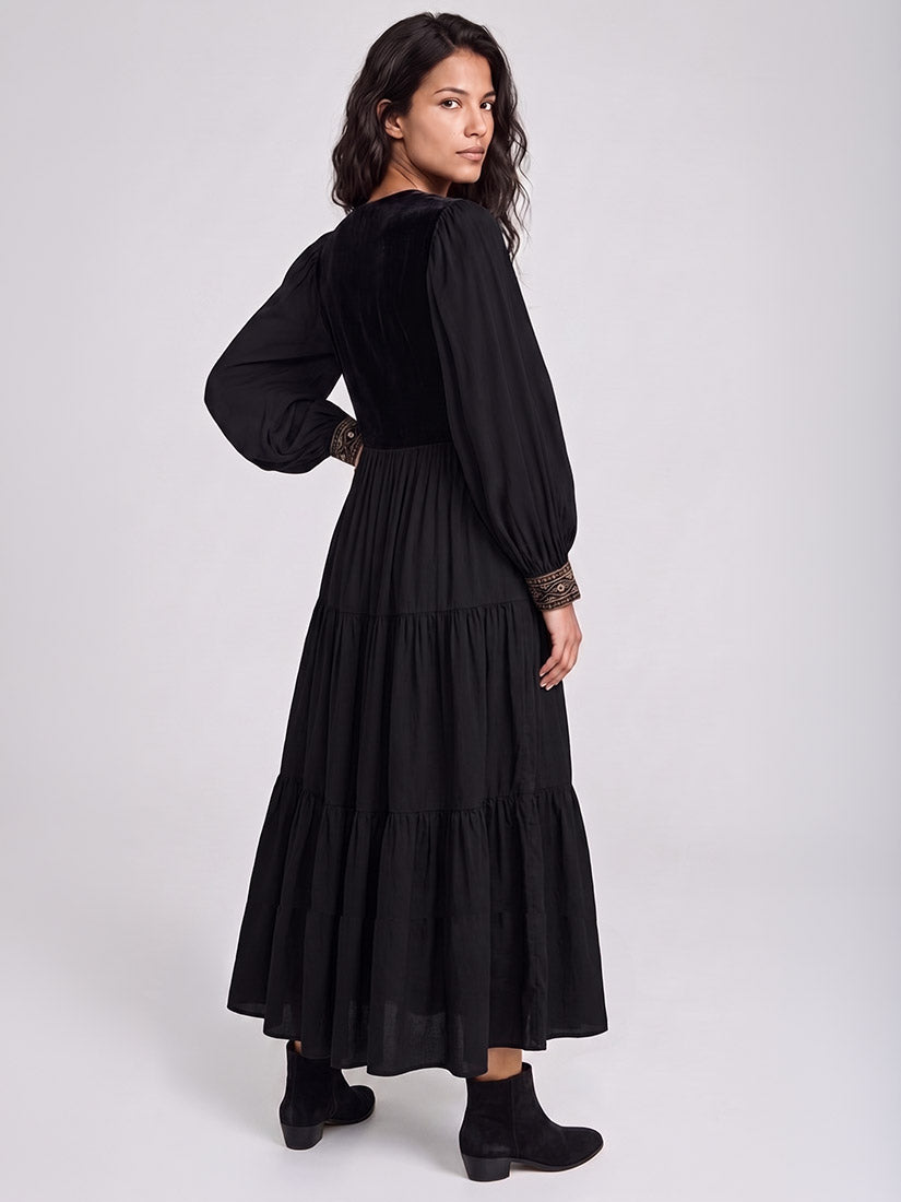 A woman modeling a long black tiered dress with a velvet-like bodice and lace cuffs, shown from the back against a light gray studio background.