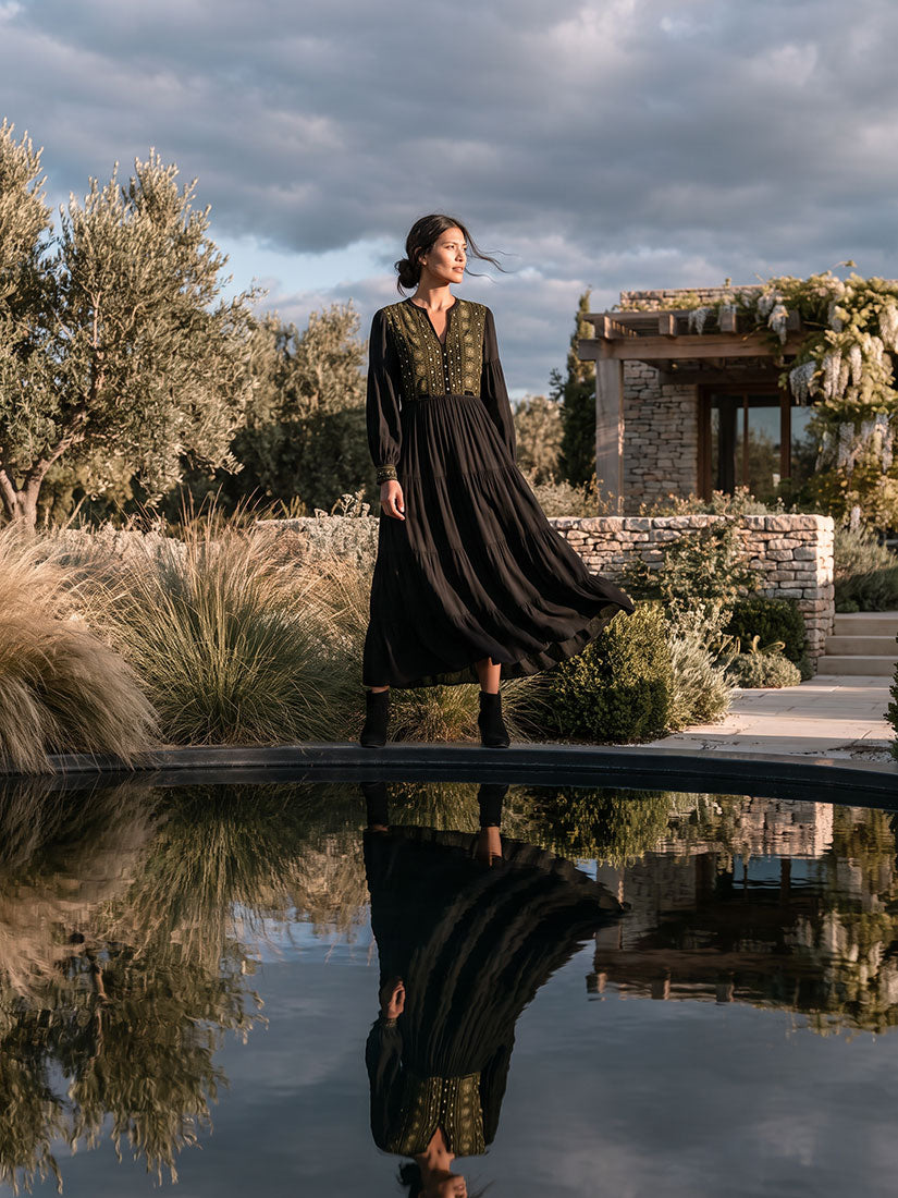 dark haired woman  in black emabroidered maison hotel dress standing over a dark reflecting pool
