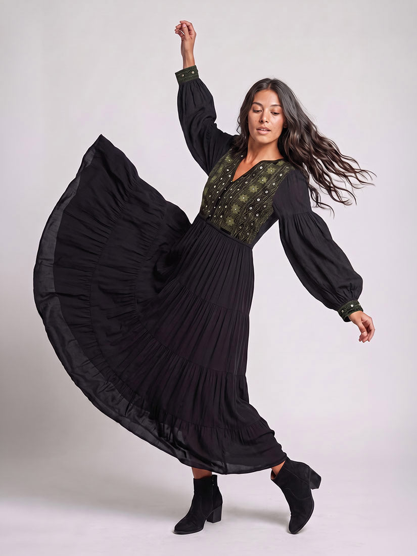 A woman wearing a long black pleated maxi dress with a dark green embroidered bodice and puff sleeves, captured mid-spin to show off the flowing skirt, against a light gray studio background.