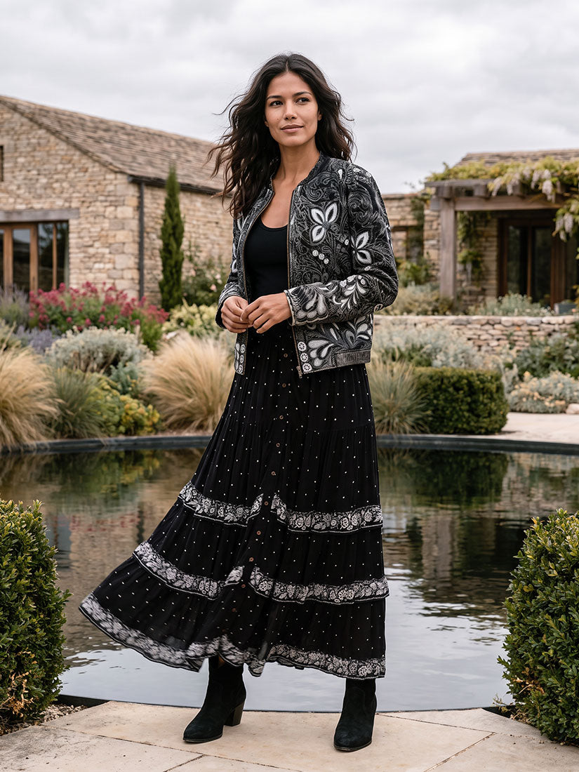 A woman stands outdoors by a stone wall and reflecting pool, wearing a long black tiered skirt with white floral lace trim, a black top, and a black embroidered jacket with white floral detailing.