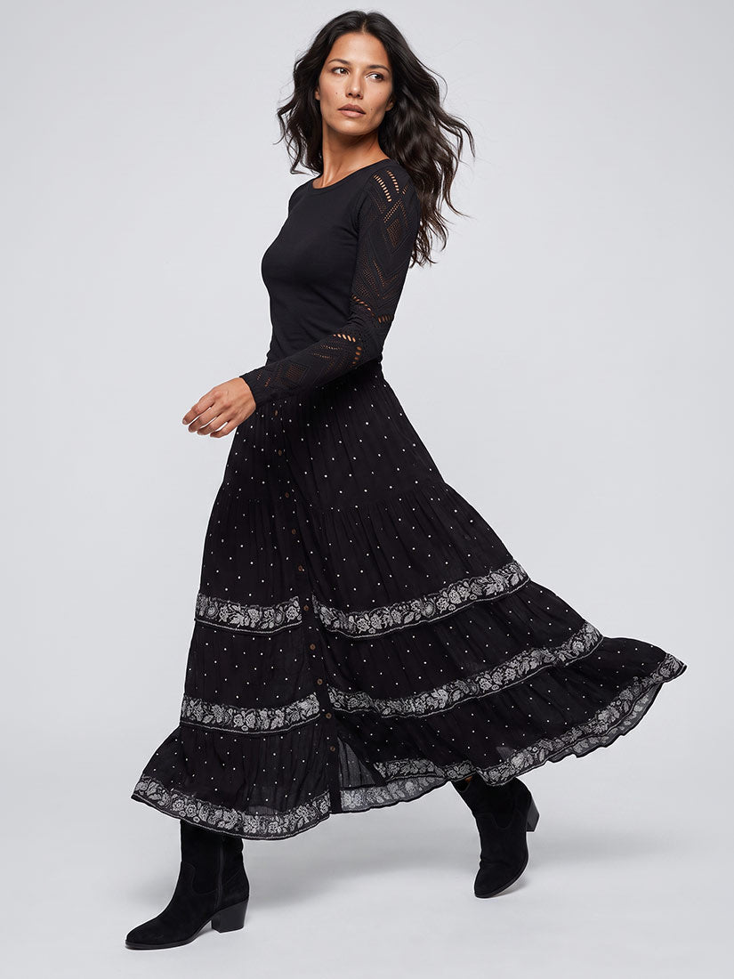 A woman modeling a long black maxi dress with sheer lace sleeves and a tiered skirt featuring small white polka dots and lace trim, paired with black ankle boots, against a light gray studio background.